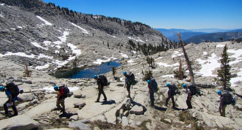 A group of people wearing helmets move across rocky terrain. There is a small body of water and mountains in the background. 